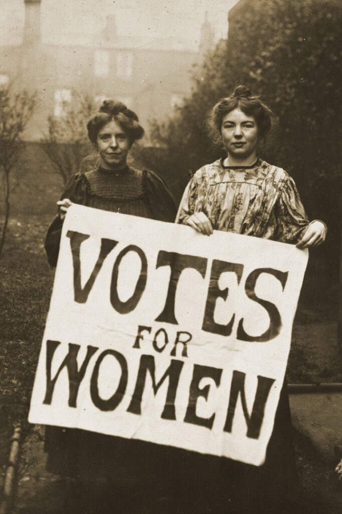 Annie Kenney and Christabel Pankhurst hold up a sign advocating for 'votes for women.' 